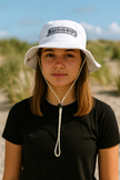 Person wearing a white bucket hat with 'Marshin Whips' logo on a beach.